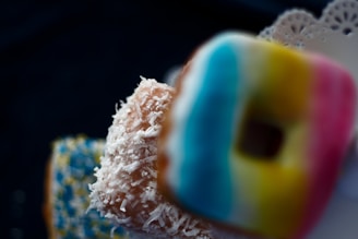 Three donuts are arranged with different toppings. One has a colorful striped glaze of blue, yellow, and pink. Another is covered with shredded coconut, giving it a textured appearance. The third donut is decorated with a combination of blue and yellow sprinkles.