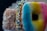 Three doughnuts are stacked closely together. The front doughnut features a gradient glaze of blue, yellow, and pink. The doughnut behind it is topped with colorful sprinkles in blue, yellow, and white, while the third doughnut in the background is covered in white coconut flakes.