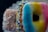 Three doughnuts are stacked closely together. The front doughnut features a gradient glaze of blue, yellow, and pink. The doughnut behind it is topped with colorful sprinkles in blue, yellow, and white, while the third doughnut in the background is covered in white coconut flakes.