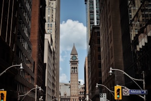 A narrow city street flanked by tall buildings converging towards a central clock tower under a partly cloudy sky. Street lamps and traffic signals are visible, with a street sign indicating Adelaide St W.