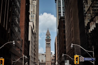 A narrow city street flanked by tall buildings converging towards a central clock tower under a partly cloudy sky. Street lamps and traffic signals are visible, with a street sign indicating Adelaide St W.