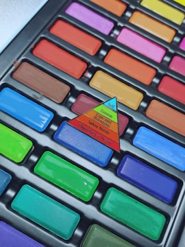 A close-up of a colorful set of rectangular pastel chalks arranged in a plastic tray. A small triangular paper pyramid illustrating Maslow's Hierarchy of Needs is placed on top of the chalks.