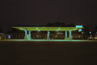 A deserted gas station at night, illuminated by greenish-yellow fluorescent lights. The station's structure and pumps are highlighted against the dark surrounding environment. The branding on the station reads 'UNO'. A dimly lit building and a lone traffic cone can be seen in the background.