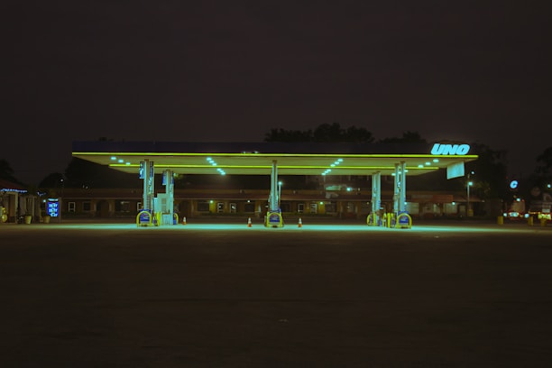 A deserted gas station at night, illuminated by greenish-yellow fluorescent lights. The station's structure and pumps are highlighted against the dark surrounding environment. The branding on the station reads 'UNO'. A dimly lit building and a lone traffic cone can be seen in the background.