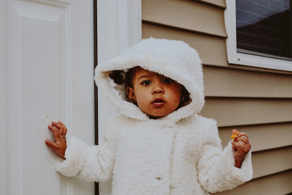 child in white winter coat standing beside white wooden door