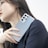 A person with long dark hair holding a sleek, modern smartphone. The phone is metallic with several camera lenses visible. The individual is dressed in a black outfit, and their gaze is directed away from the camera.