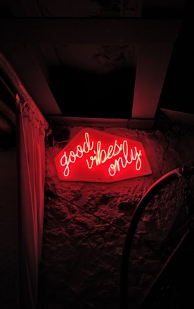 A red neon sign with the words 'good vibes only' is mounted on a textured stone or brick wall. The lighting creates a warm and inviting glow, contrasting against the dark surroundings.