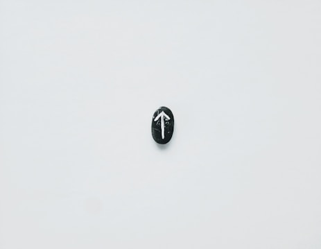 A single black stone is centered on a plain white background. The stone features a white arrow pointing upwards, painted on its surface.