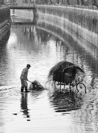 A person wearing boots stands in the middle of a shallow canal, holding a rake and gathering grass or debris into a pile. Nearby, a bicycle with a large basket, also filled with grass, is partially submerged in the water. The setting appears to be urban, with a bridge crossing the canal in the background and foliage lining the banks.