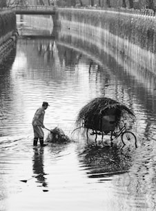 A person wearing boots stands in the middle of a shallow canal, holding a rake and gathering grass or debris into a pile. Nearby, a bicycle with a large basket, also filled with grass, is partially submerged in the water. The setting appears to be urban, with a bridge crossing the canal in the background and foliage lining the banks.