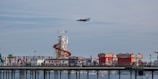 A coastal amusement park with a prominent 'Family Fun Park' sign is situated on a pier extending over the water. The park features a helter-skelter slide with a red spiral ramp and various colorful stalls and attractions. A vintage airplane is flying overhead against a backdrop of a partly cloudy sky.