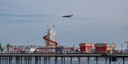A coastal amusement park with a prominent 'Family Fun Park' sign is situated on a pier extending over the water. The park features a helter-skelter slide with a red spiral ramp and various colorful stalls and attractions. A vintage airplane is flying overhead against a backdrop of a partly cloudy sky.