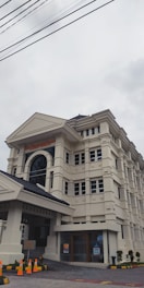 A large, multi-story building with classical architectural elements, such as columns and arches, featuring cream-colored walls and a dark, tiled roof. Orange traffic cones are placed around the entrance area, and there are a few small potted plants near the entrance. Overhead power lines are visible against the cloudy sky.