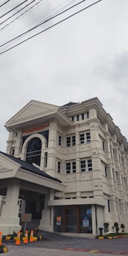 A large, multi-story building with classical architectural elements, such as columns and arches, featuring cream-colored walls and a dark, tiled roof. Orange traffic cones are placed around the entrance area, and there are a few small potted plants near the entrance. Overhead power lines are visible against the cloudy sky.