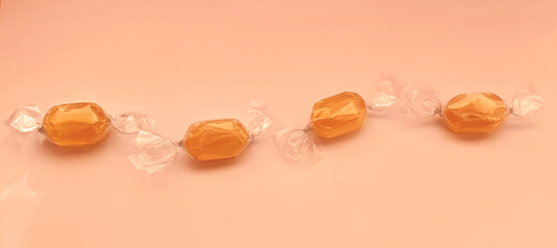 Four amber caramels in clear twist wrappers laid in a row on a pink background