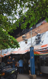 A street market scene with a group of people gathered outside a shop. The shop has colorful banners with various advertisements. There is a vintage-style street lamp post in front. The scene is shaded by tree branches laden with green leaves, contributing to a lively and bustling atmosphere.