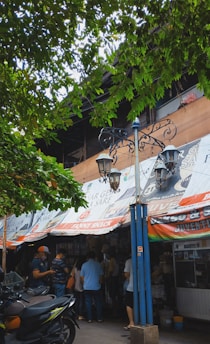 A street market scene with a group of people gathered outside a shop. The shop has colorful banners with various advertisements. There is a vintage-style street lamp post in front. The scene is shaded by tree branches laden with green leaves, contributing to a lively and bustling atmosphere.