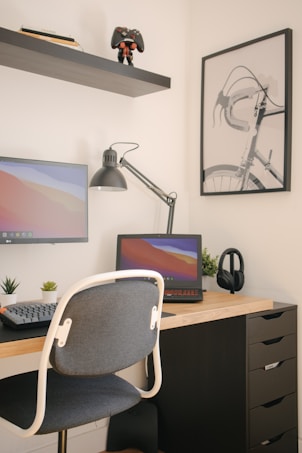 A modern workspace with a wooden desk featuring a laptop and a monitor displaying a colorful gradient. A desk lamp is positioned towards the center, with potted plants adding greenery. Headphones rest subtly on the desk. Above, a shelf holds books and a toy figurine, providing a playful touch. A minimalist bicycle poster adorns the wall.