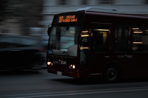 A bus moves swiftly through an urban setting, showing motion blur as it travels along the road. The destination sign on the bus is illuminated, and its headlights are on. The background is dark, with little detail visible, suggesting it's either evening or a dimly lit area.