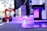 A high-tech laser cutter or CNC machine is in operation, focusing a beam onto a reflective surface. The machine is surrounded by a backdrop of blurred industrial elements, with visible purple and blue lighting highlighting the setup.