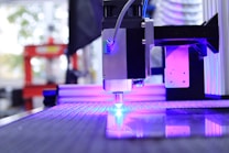 A high-tech laser cutter or CNC machine is in operation, focusing a beam onto a reflective surface. The machine is surrounded by a backdrop of blurred industrial elements, with visible purple and blue lighting highlighting the setup.