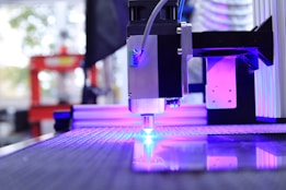 A high-tech laser cutter or CNC machine is in operation, focusing a beam onto a reflective surface. The machine is surrounded by a backdrop of blurred industrial elements, with visible purple and blue lighting highlighting the setup.