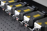 A row of industrial laser devices with metallic casings and warning labels, mounted on a perforated metal surface. Each device has a sleek design with a protruding component and visible screws.