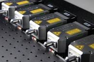A row of industrial laser devices with metallic casings and warning labels, mounted on a perforated metal surface. Each device has a sleek design with a protruding component and visible screws.