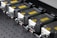 A row of industrial laser devices with metallic casings and warning labels, mounted on a perforated metal surface. Each device has a sleek design with a protruding component and visible screws.