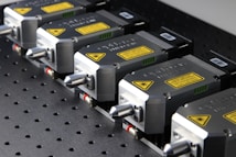 A row of industrial laser devices with metallic casings and warning labels, mounted on a perforated metal surface. Each device has a sleek design with a protruding component and visible screws.