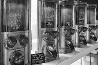 A row of vintage-style coffee bean dispensers with hand-written labels indicating different blends and variants. The dispensers have metal finishes and prominent dispensing spouts. A small sign on the shelf mentions coffee beans for sale ground or whole.