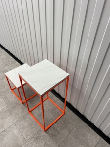 Two modern, minimalist square tables with bright orange metal frames and white marble tops are placed on a tiled floor next to a metal wall with a vertical striped pattern.