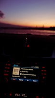 A car dashboard with an infotainment screen displaying music information, showing a song by Damien Rice during twilight. The background features a serene sunset over a body of water, with a gradient of colors from orange to purple in the sky.