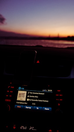 A car dashboard with an infotainment screen displaying music information, showing a song by Damien Rice during twilight. The background features a serene sunset over a body of water, with a gradient of colors from orange to purple in the sky.