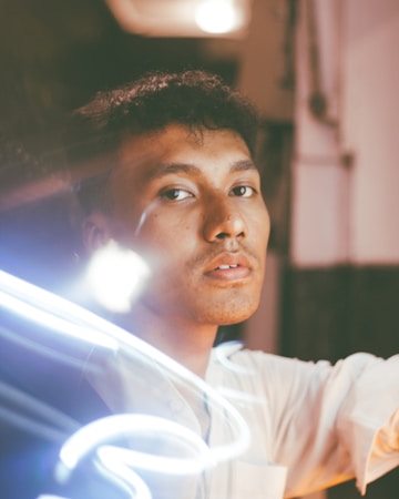 A young man with curly hair gazes into the camera with a neutral expression. The scene is illuminated by a swirl of light trails that create a dynamic composition. The background is slightly out of focus, with warm tones and soft lighting.