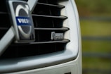 Close-up of a car grille featuring the Volvo logo and an R-Design badge. The image focuses on the sleek design and branding of the vehicle, with a blurred green background.