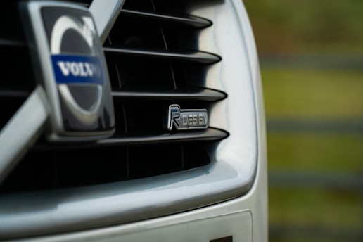 Close-up of a car grille featuring the Volvo logo and an R-Design badge. The image focuses on the sleek design and branding of the vehicle, with a blurred green background.