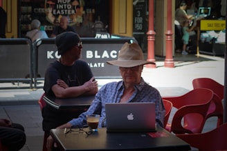 An elderly man wearing a straw hat is sitting at an outdoor café table, working on a laptop with a cup of coffee in front of him. He is wearing a blue plaid shirt and glasses. Another man in black clothing and a beanie is sitting at the adjacent table. Behind them, there are people walking and shopping, with store signs and advertisements visible in the background.
