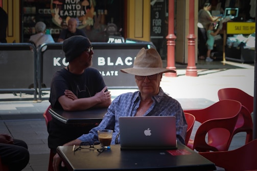An elderly man wearing a straw hat is sitting at an outdoor café table, working on a laptop with a cup of coffee in front of him. He is wearing a blue plaid shirt and glasses. Another man in black clothing and a beanie is sitting at the adjacent table. Behind them, there are people walking and shopping, with store signs and advertisements visible in the background.