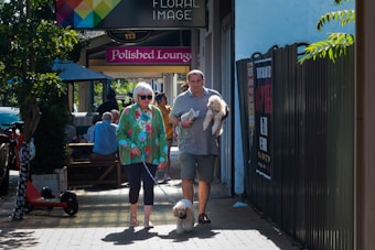 A couple walks down a sidewalk on a sunny day. The woman, wearing a colorful shirt and sunglasses, is leading a small white dog on a leash. The man beside her is carrying another small white dog. They pass by a caf&eacute; where people are sitting under large umbrellas at wooden tables. Banners and signs are visible on a fence to the right.