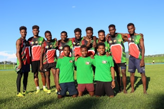 A group of young men stand on a grassy field, wearing sports jerseys. Most of them are dressed in black, red, and green athletic gear, while two in the front wear green shirts and shorts. They are smiling and appear relaxed, with some of them having their arms around each other's shoulders.