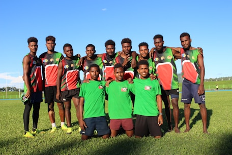A group of young men stand on a grassy field, wearing sports jerseys. Most of them are dressed in black, red, and green athletic gear, while two in the front wear green shirts and shorts. They are smiling and appear relaxed, with some of them having their arms around each other's shoulders.