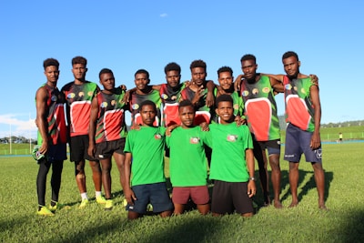 A group of young men stand on a grassy field, wearing sports jerseys. Most of them are dressed in black, red, and green athletic gear, while two in the front wear green shirts and shorts. They are smiling and appear relaxed, with some of them having their arms around each other's shoulders.