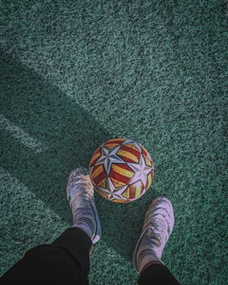 Two shoes, covered in dust, stand on either side of a colorful soccer ball on a green synthetic field.