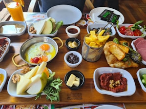 A variety of breakfast foods are neatly arranged on a wooden table. The items include a pan of eggs sunny side up with cherry tomatoes, a basket of golden French fries, slices of cheese, butter, chocolate spread, nuts, and fruit slices. There are also plates of vegetables such as tomatoes, cucumbers, and leafy greens, as well as a glass of orange juice. In addition, there are plates with red paste topped with nuts, cooked meat, and a stack of green peppers with potatoes. Empty plates and utensils are placed in the background.