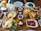 A variety of breakfast foods are neatly arranged on a wooden table. The items include a pan of eggs sunny side up with cherry tomatoes, a basket of golden French fries, slices of cheese, butter, chocolate spread, nuts, and fruit slices. There are also plates of vegetables such as tomatoes, cucumbers, and leafy greens, as well as a glass of orange juice. In addition, there are plates with red paste topped with nuts, cooked meat, and a stack of green peppers with potatoes. Empty plates and utensils are placed in the background.