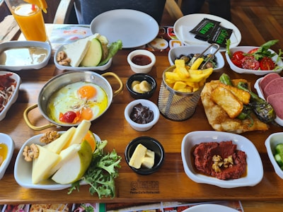 A variety of breakfast foods are neatly arranged on a wooden table. The items include a pan of eggs sunny side up with cherry tomatoes, a basket of golden French fries, slices of cheese, butter, chocolate spread, nuts, and fruit slices. There are also plates of vegetables such as tomatoes, cucumbers, and leafy greens, as well as a glass of orange juice. In addition, there are plates with red paste topped with nuts, cooked meat, and a stack of green peppers with potatoes. Empty plates and utensils are placed in the background.
