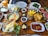 A variety of breakfast foods are neatly arranged on a wooden table. The items include a pan of eggs sunny side up with cherry tomatoes, a basket of golden French fries, slices of cheese, butter, chocolate spread, nuts, and fruit slices. There are also plates of vegetables such as tomatoes, cucumbers, and leafy greens, as well as a glass of orange juice. In addition, there are plates with red paste topped with nuts, cooked meat, and a stack of green peppers with potatoes. Empty plates and utensils are placed in the background.
