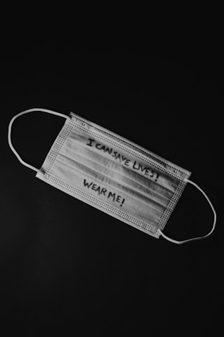 A disposable face mask lies against a dark background. The mask has handwritten text on it that reads 'I CAN SAVE LIVES! WEAR ME!'.