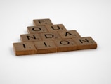 Wooden Scrabble tiles are arranged to spell the word 'FOUNDATION' in a pyramid shape on a plain white background.